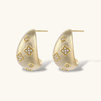Curved gold-plated hoop earrings with four-leaf clover and diamond-shaped crystal details on white background