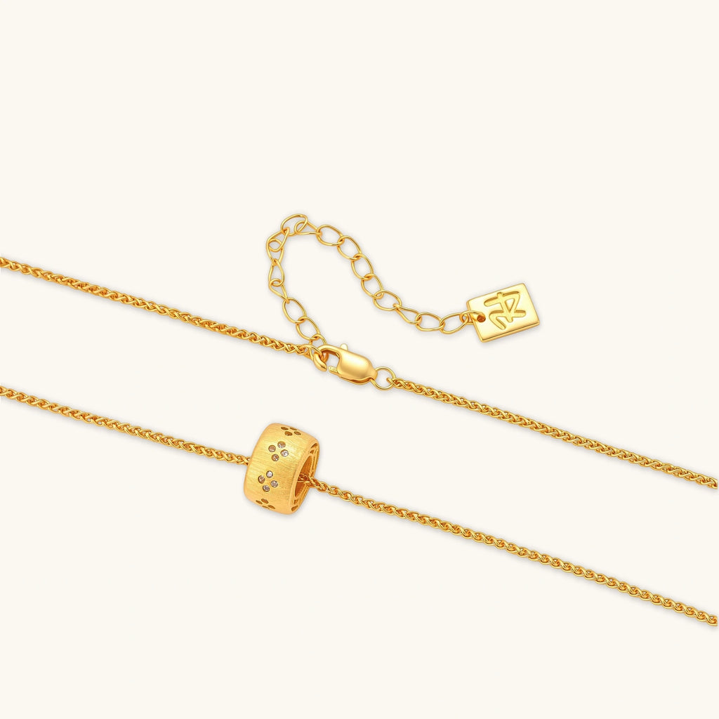 Gold-plated cylindrical ring pendant with brushed finish and scattered crystal accents on a wheat-link chain, cream background.