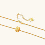 Gold-plated cylindrical ring pendant with brushed finish and scattered crystal accents on a wheat-link chain, cream background.