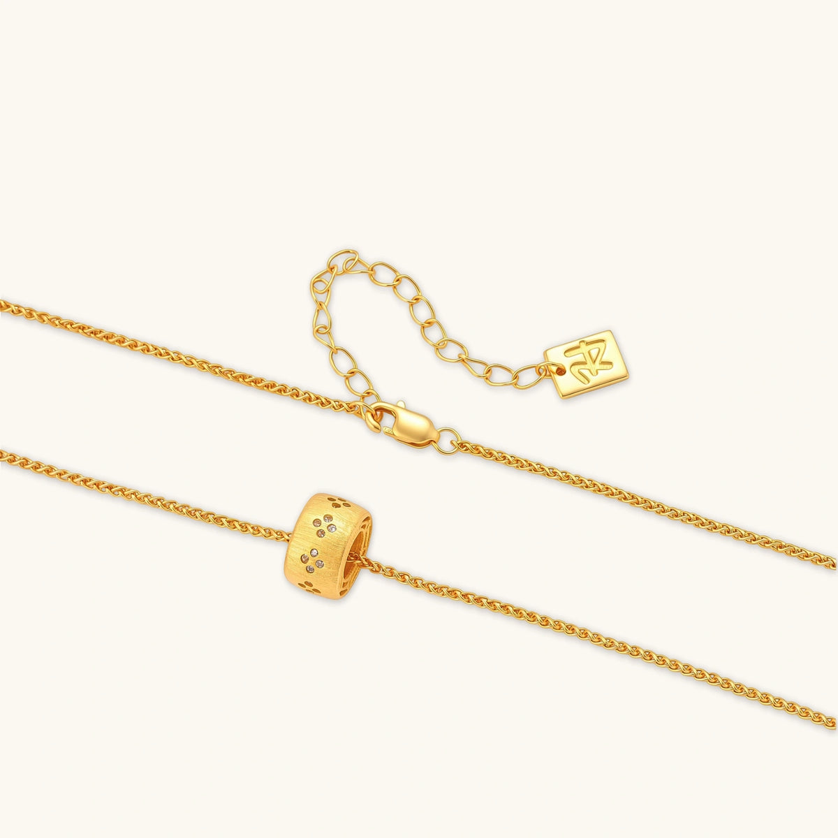 Gold-plated cylindrical ring pendant with brushed finish and scattered crystal accents on a wheat-link chain, cream background.